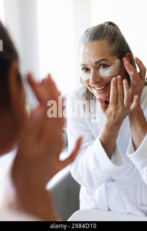 In bagno a casa, donna caucasica matura che applica maschera per gli occhi, guardando nello specchio Foto Stock