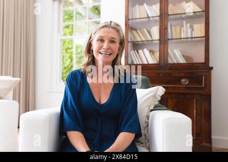 Una donna caucasica senior sorridente durante una videochiamata a casa, con indosso un vestito blu Foto Stock