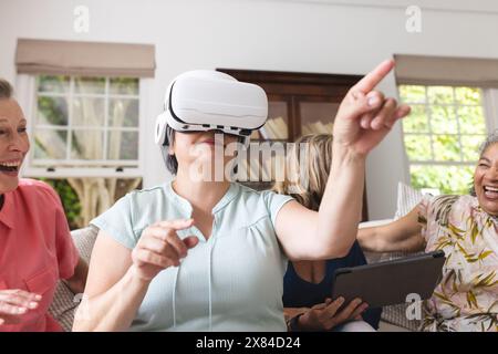Diverse amiche anziane che ridono a casa, una che punta con la cuffia Foto Stock