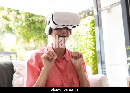 Una donna caucasica anziana che indossa un visore per la realtà virtuale, ridendo a casa Foto Stock