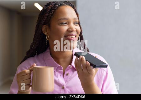 In un ufficio moderno per affari, una giovane donna birazziale sorride, tenendo in mano uno smartphone e una tazza Foto Stock