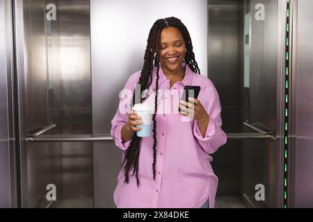 Nel moderno ufficio per affari, giovane donna birazziale in ascensore, che tiene in mano una tazza di caffè Foto Stock