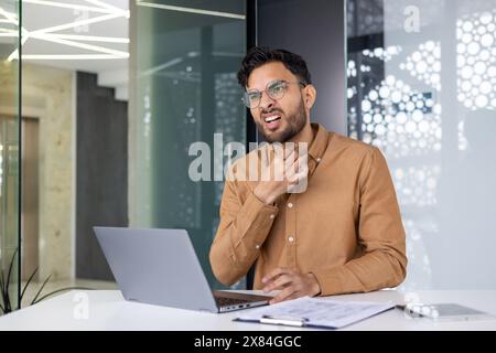 Uomo con una camicia marrone seduto a una scrivania con un portatile, toccando la gola con disagio, indicando dolore o malattia. Foto Stock