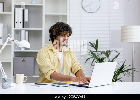 Giovane uomo con auricolare che lavora su un computer portatile alla scrivania dell'ufficio, creando un ambiente di lavoro produttivo e mirato in un ufficio moderno e luminoso. Foto Stock