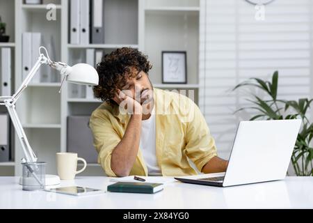 Un uomo stanco che dorme alla scrivania con un notebook, sentendosi esausto in un ufficio domestico. Foto Stock