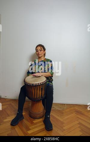 Una giovane donna latina siede su uno sgabello suonando un tamburo yembe a casa. Il tamburo è in legno e presenta un design nero e marrone. La donna indossa un shi blu Foto Stock