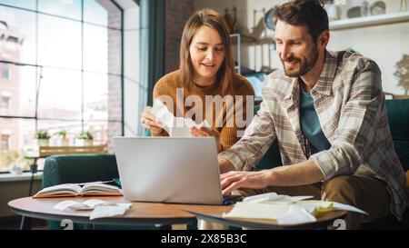 Fare contabilità a casa: Coppia felice che usa il computer portatile e si siede sul divano dell'appartamento. Young Family che compila moduli fiscali, documenti ipotecari, bollette, assegni, saldi, le fatture sono in ordine Foto Stock