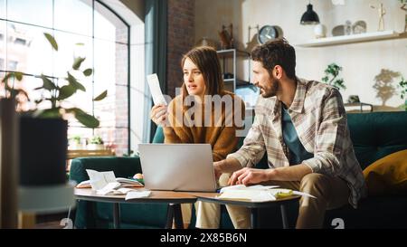 Fare contabilità a casa: Coppia felice che usa il computer portatile e si siede sul divano dell'appartamento. Young Family che compila moduli fiscali, documenti ipotecari, bollette, assegni, saldi, le fatture sono in ordine Foto Stock