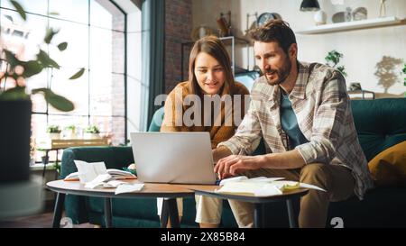 Fare contabilità a casa: Coppia felice che usa il computer portatile e si siede sul divano dell'appartamento. Young Family che compila moduli fiscali, documenti ipotecari, bollette, assegni, saldi, le fatture sono in ordine Foto Stock