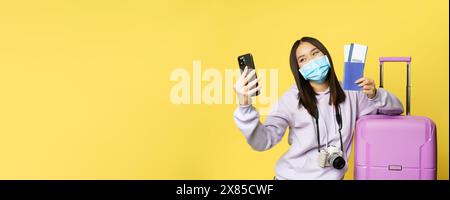 Felice donna coreana che prende selfie con passaporto e biglietti di volo, in vacanza, in piedi vicino alla grande carina valigia rosa, sfondo giallo Foto Stock