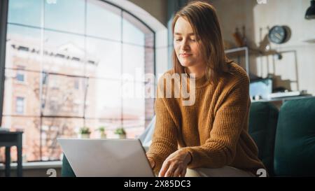 La giovane donna sorridente che lavora da casa sul computer portatile nel suo appartamento Sunny Cozy si rilassa e si appoggia su un divano. Imprenditrice creativa di successo felice dopo aver finito il lavoro remoto Foto Stock