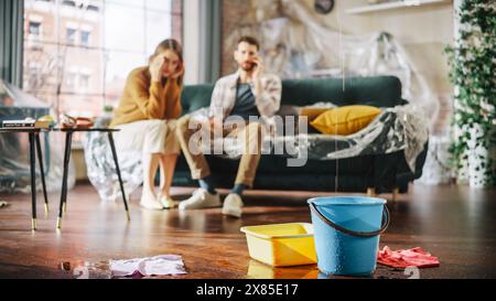 Il tetto perde o il tubo si rompe a casa: Una coppia disperata seduta su un divano che guarda come l'acqua gocciola nei secchi nel loro soggiorno. Catastrofe, distaster e rovina finanziaria Foto Stock