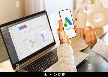 Uomo che utilizza un telefono cellulare con Google Ads sullo schermo e in background un computer che mostra le statistiche degli annunci. Rosario, Argentina - 23 marzo 2024 Foto Stock