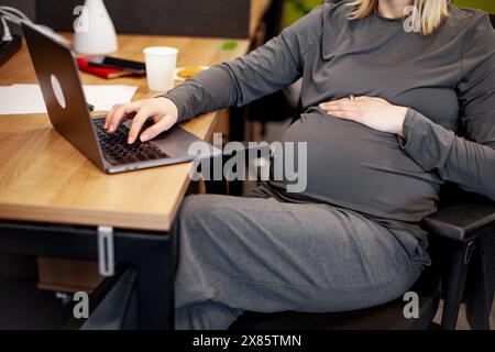 Primo piano di una donna incinta che lavora su un notebook. Donna d'affari incinta corta che scrive sul computer portatile mentre è seduta sul posto di lavoro in ufficio Foto Stock