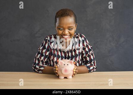 Una donna afroamericana con un aspetto pazzo e un sorriso guarda un salvadanaio a forma di maiale. Foto Stock