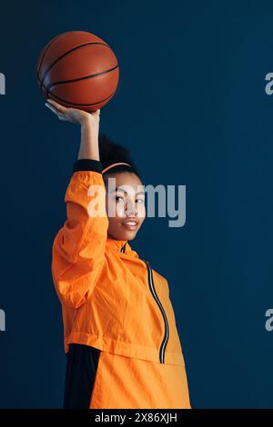 Giovane giocatrice di basket che tiene un pallacanestro su un braccio teso. Ritratto in studio di una giocatrice di basket con abbigliamento sportivo arancione. Foto Stock