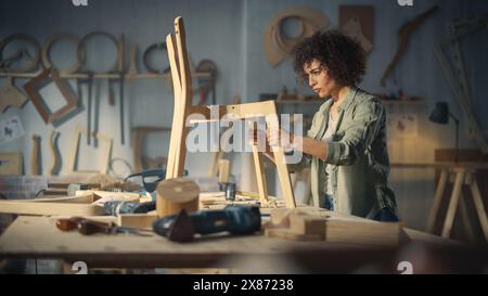 Ritratto di un giovane carpentiere che controlla un Blueprint e inizia a montare una sedia di legno. Designer di mobili professionale che lavora in uno studio in un loft con strumenti sulle pareti. Foto Stock