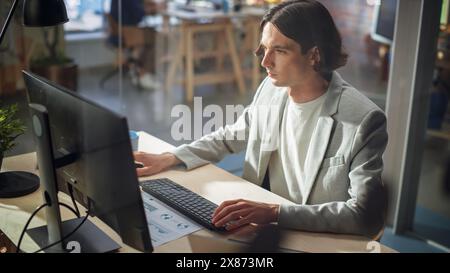 Elegante manager dai capelli lunghi che indossa una giacca grigia e lavora da una scrivania in ufficio creativo. Young Handsome Man Using Desktop computer in Marketing Agency. Colleghi che lavorano in background. Foto Stock