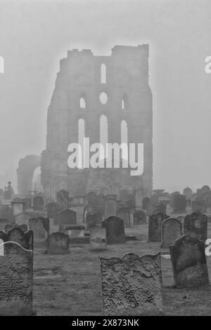 Una scena nebbiosa di un vecchio cimitero con numerose lapidi in primo piano. Sullo sfondo, le rovine di un grande, antico edificio in pietra Foto Stock