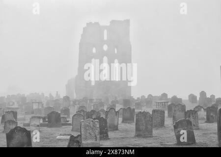 Un cimitero nebbioso con antiche lapidi e le rovine di un grande edificio sullo sfondo. Foto Stock