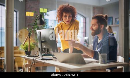 Manager e Specialista donna lavorano su un progetto dietro una scrivania con schermo di un computer e un notebook. I colleghi lavorano in un Creative Loft Office e discutono di una nuova strategia pubblicitaria per un cliente. Foto Stock