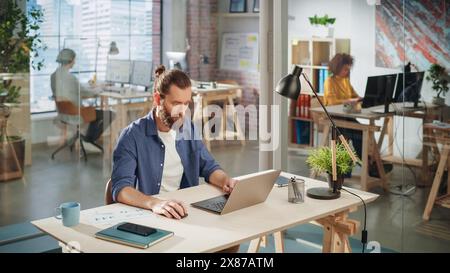 Bellissimo manager con capelli lunghi seduto a una scrivania in Creative Office. Giovane uomo alla moda che utilizza un computer portatile presso l'agenzia di marketing. Colleghi che lavorano in background. Vista ad angolo alto. Foto Stock