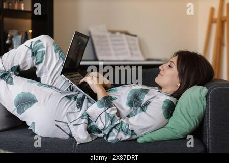 Donna d'affari rilassata in pigiama che utilizza un computer portatile sdraiato sul divano e lavora a casa Foto Stock