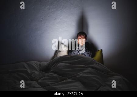 Uomo depresso che copre il viso con le mani sedute sul letto in una stanza buia Foto Stock