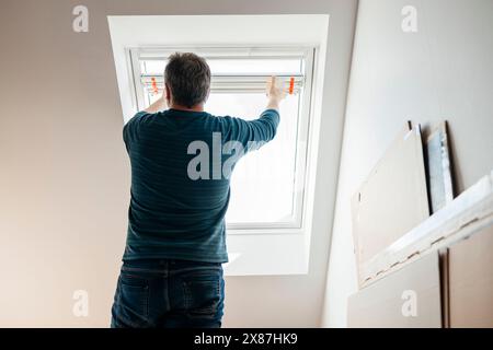 Uomo che installa tende sulla finestra dell'attico a casa Foto Stock