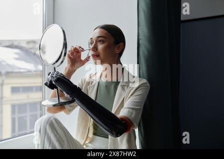 Giovane donna con braccio bionico che guarda se stessa allo specchio usando un arricciacapelli ciglia Foto Stock