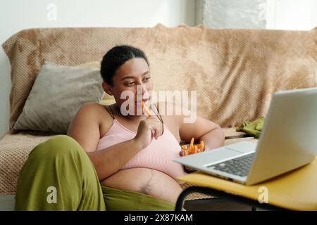 Donna incinta che guarda il portatile e mangia carote seduto nel soggiorno di casa Foto Stock