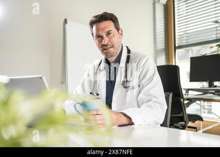 Medico sicuro seduto con una tazza di caffè e un computer portatile alla scrivania dell'ufficio Foto Stock