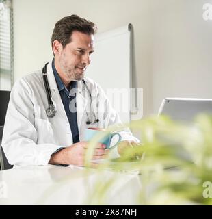 Medico sicuro seduto con una tazza di caffè utilizzando un computer portatile alla scrivania dell'ufficio Foto Stock