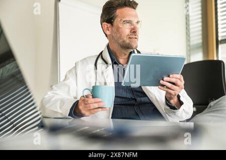 Medico che indossa occhiali intelligenti utilizzando un tablet PC seduto alla scrivania dell'ufficio in clinica Foto Stock