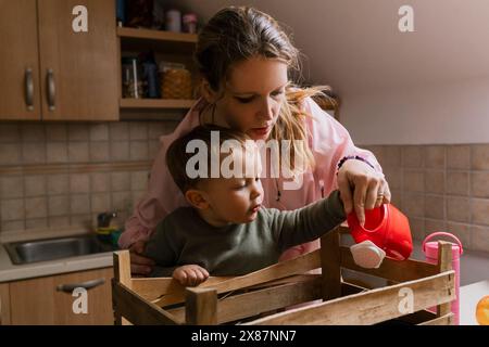 Madre e figlio annaffiano le piantine in una cassa di legno in cucina Foto Stock