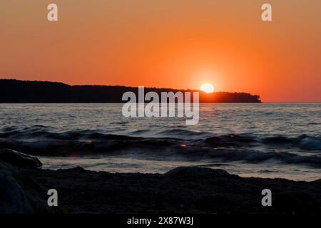 Il sole sta lentamente affondando sotto l'orizzonte, gettando un caldo bagliore sul tranquillo corpo d'acqua. Il cielo è dipinto con sfumature di rosso e di arancia Foto Stock