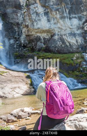 Una donna è seduta su una roccia vicino a una cascata, mentre scatta una foto con la sua macchina fotografica Foto Stock