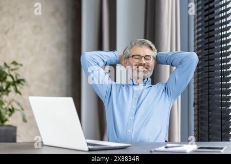 Un uomo dai capelli grigi si prende una pausa alla scrivania del suo ufficio, sorridendo e rilassato con un notebook Foto Stock