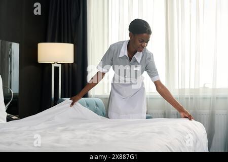 Ritratto minimale di una giovane donna afroamericana sorridente come governante che indossa l'uniforme che fa il letto nello spazio fotocopie della camera d'albergo Foto Stock