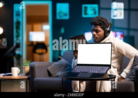 Uomo afroamericano che si prende una pausa dal lavoro per ascoltare musica e leggere dal tablet, concentrarsi sul notebook mockup. Telelavoratore che indossa le cuffie per divertirsi accanto a un notebook con schermo isolato Foto Stock