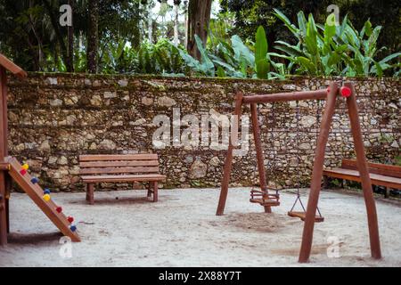 Tranquillo parco giochi per bambini immerso in un parco tropicale, con panchina in legno, altalena e colorata struttura per arrampicate. Vegetazione lussureggiante e un histo Foto Stock