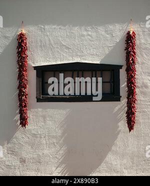 Illuminazione laterale dei ristras rossi di peperoncino appesi su pareti bianche con finestre in legno nero all'esterno della tradizionale dimora del New Mexico meridionale a Mesilla, NEW MEXICO Foto Stock