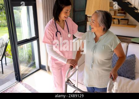 A casa, giovane infermiera asiatica e paziente di mezza età che usa il walker, entrambi sorridenti Foto Stock