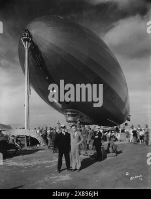 Uomo d'affari con la tuta e cravatta in posa con il braccio intorno al capitano del Graf Zeppelin mentre una folla di uomini e donne si alzano in piedi e osservano. Il Graf Zeppelin è in modalità tiedown a terra Foto Stock