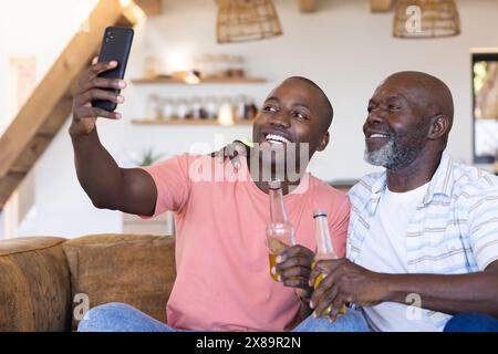 Padre afroamericano anziano, figlio adulto a casa con bevande e selfie Foto Stock