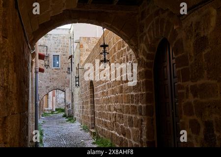 Uno stretto passaggio in pietra conduce attraverso un vicolo lastricato di ciottoli. Pareti con archi e lanterne creano un'atmosfera rustica, la città vecchia di Rodi, Foto Stock