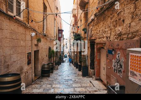 Un uomo passeggia lungo una pittoresca strada di pietra fiancheggiata da verde e botti di legno a la Valletta, Malta Foto Stock