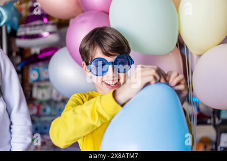 ragazzo con occhiali da festa blu che tiene in mano il palloncino blu e sorride Foto Stock