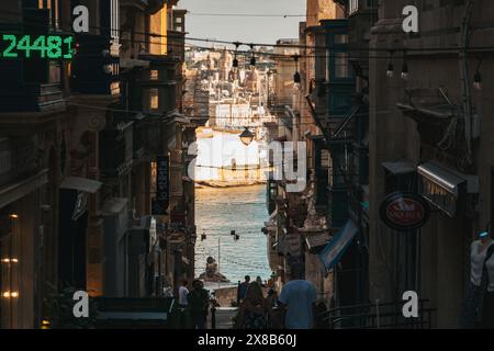 Una strada stretta a la Valletta, Malta, fiancheggiata da edifici e vetrine. La strada conduce ad una vista del porto al tramonto Foto Stock