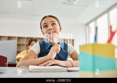 Una giovane ragazza seduta a una scrivania, profondamente pensata, tiene in mano una penna, circondata da una vivace aula Foto Stock